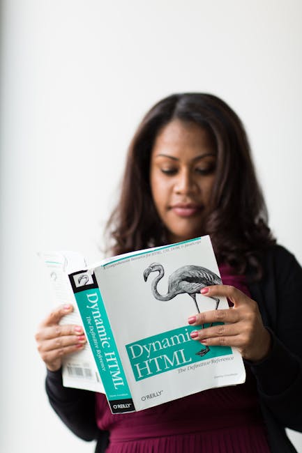 A woman with dark, wavy hair and light skin, dressed in a black blazer over a dark pink top, is standing indoors against a plain white wall. She is holding an open book titled 'Dynamic HTML' with a pr
